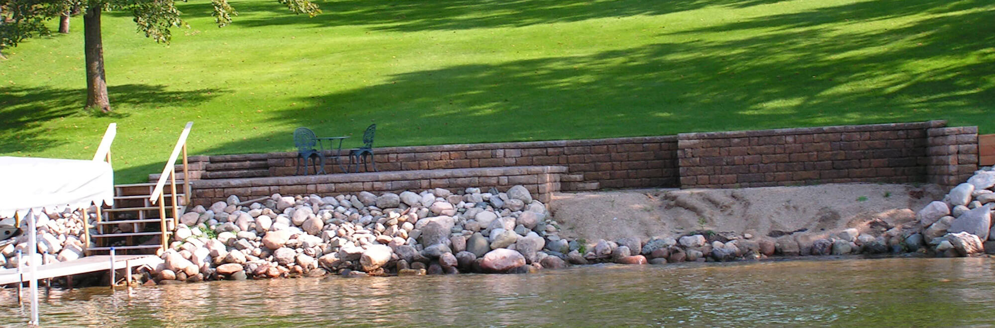 Outdoor living space created along the lakeshore using retaining wall block, patio pavers, boulders, and beach sand